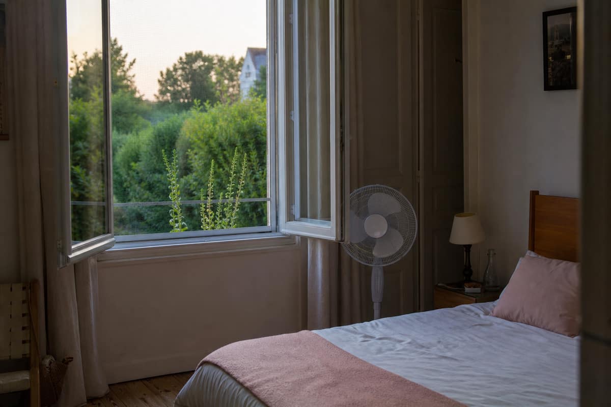Fenêtre de chambre équipée d’une moustiquaire et ventilateur près du lit
