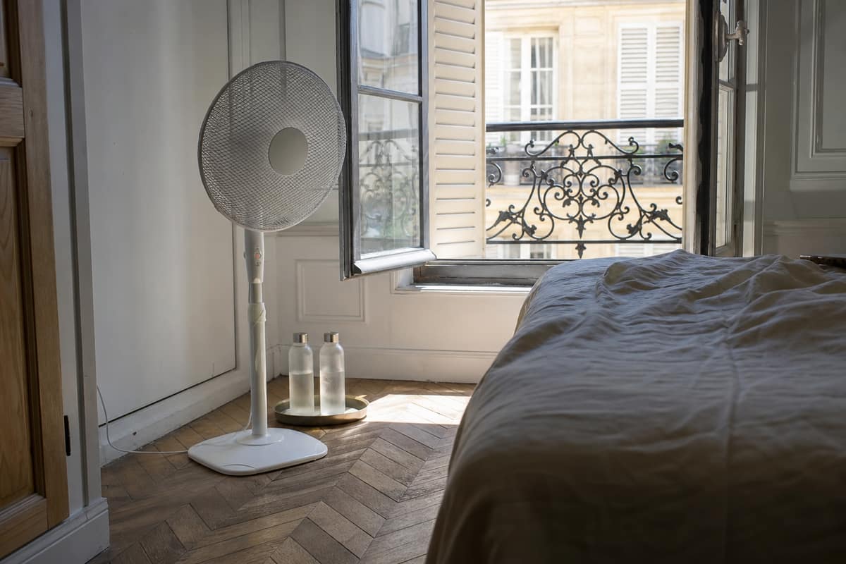 Ventilateur sur pied près d’une fenêtre avec bouteilles glacées dans un bac