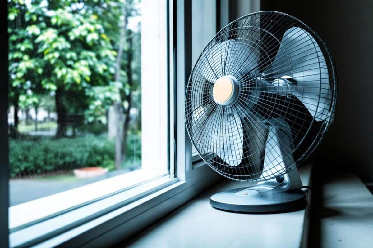 Ventilateur sur pied soufflant vers une fenêtre ouverte dans une chambre estivale
